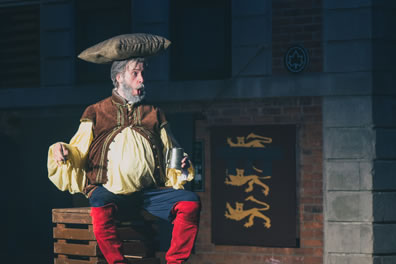 Falstaff, with protruding belly, sits on a crate and holds a tankard in his left hand, his right hand held out as if in regal posture with his mouth puckered in great pronouncement: he has a pillow on his head, and he's wearing his normal outfit of buckskin vest over yellow shirt, blue pants, and bright redk, knee-high boots. A banner of three royal lions hang on a brick wall in the background.